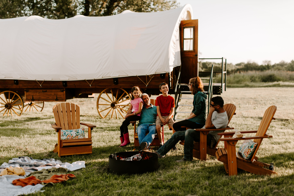 A family sits around a campfire with PlainsCraft Covered Wagons behind them. PlainsCraft offers overnight accommodations for outdoor destinations that are researching if glamping is good for a business